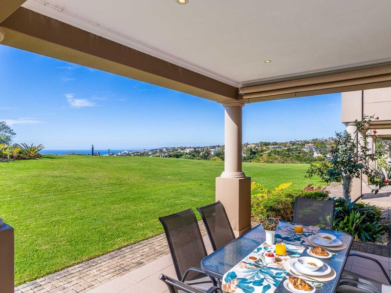 A spacious covered patio featuring paved flooring and structural columns, offering expansive views over a manicured lawn and the distant coastline.