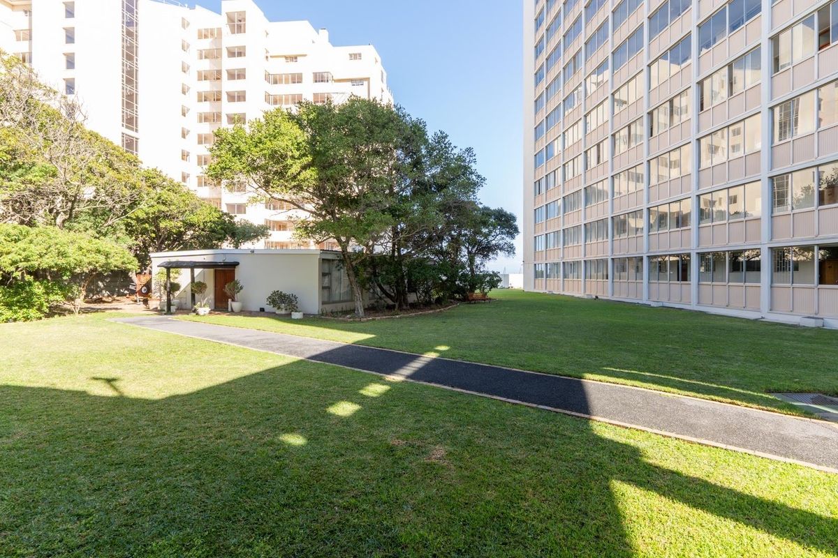 A well-maintained communal garden area featuring a paved walkway between apartment building blocks with mature trees and manicured lawn.