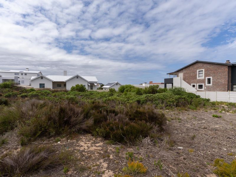 A vacant residential plot featuring natural coastal vegetation and sandy terrain, situated in a developed neighborhood with established homes.