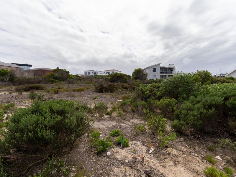 A vacant residential plot featuring native coastal vegetation and sandy terrain, situated in an established neighborhood with surrounding residential properties.