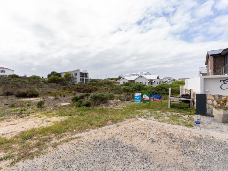 A vacant residential plot featuring natural vegetation, stone-clad boundary walling, and a paved driveway entrance in a coastal residential area.
