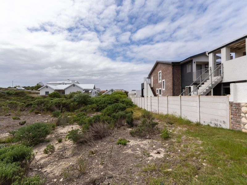 A view of the vacant residential plot showing the adjacent property's rear elevation, boundary wall, and natural coastal vegetation.