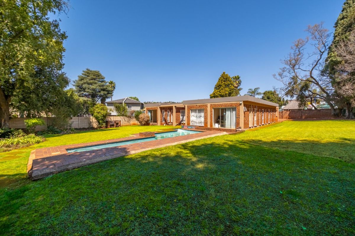 Rear view of the property featuring a rectangular swimming pool, brick paved surround, and a spacious lawn area with established trees.