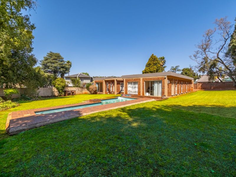 Rear view of the property featuring a rectangular swimming pool, brick paved surround, and a spacious lawn area with established trees.