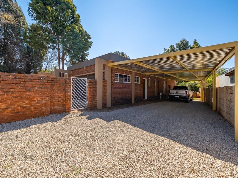Front exterior view featuring a brick boundary wall, metal gate, and a spacious covered carport over a gravel driveway.