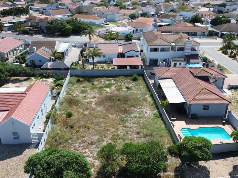 An aerial view of a vacant residential plot situated in a developed suburban neighborhood, surrounded by existing homes and boundary fencing.