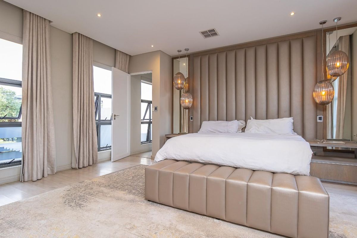 Spacious modern bedroom featuring a floor-to-ceiling upholstered headboard, integrated mirrored panels, recessed lighting, and large aluminium windows with neutral curtains.