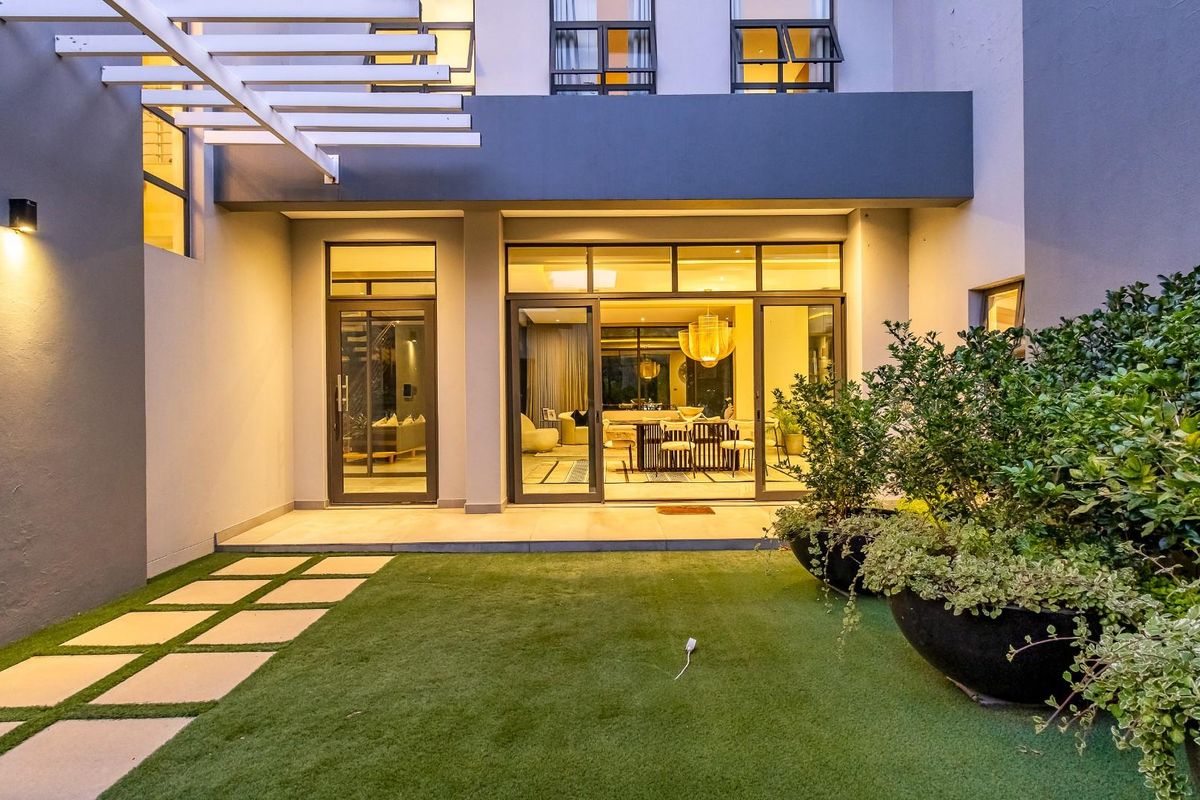 A modern rear exterior view featuring a paved patio, artificial lawn, and large sliding glass doors providing seamless indoor-outdoor flow.
