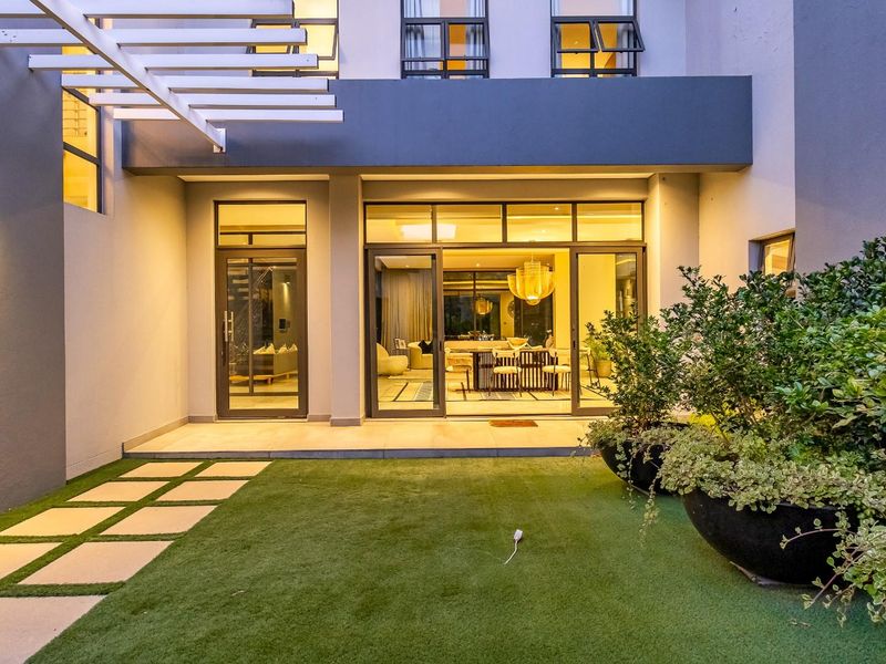 A modern rear exterior view featuring a paved patio, artificial lawn, and large sliding glass doors providing seamless indoor-outdoor flow.