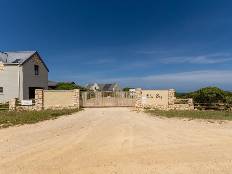 Entrance view of the Blue Bay residential estate featuring a gated access point, stone masonry pillars, and a gravel driveway.