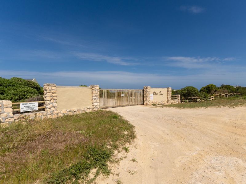 Entrance gate to Blue Bay residential area featuring stone-clad pillars and a metal security gate accessible via a sand road.