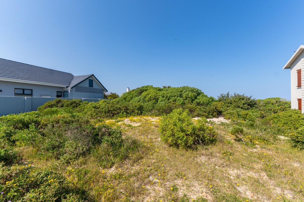 A view of a vacant residential plot featuring natural coastal vegetation and sandy terrain, situated between existing residential properties.