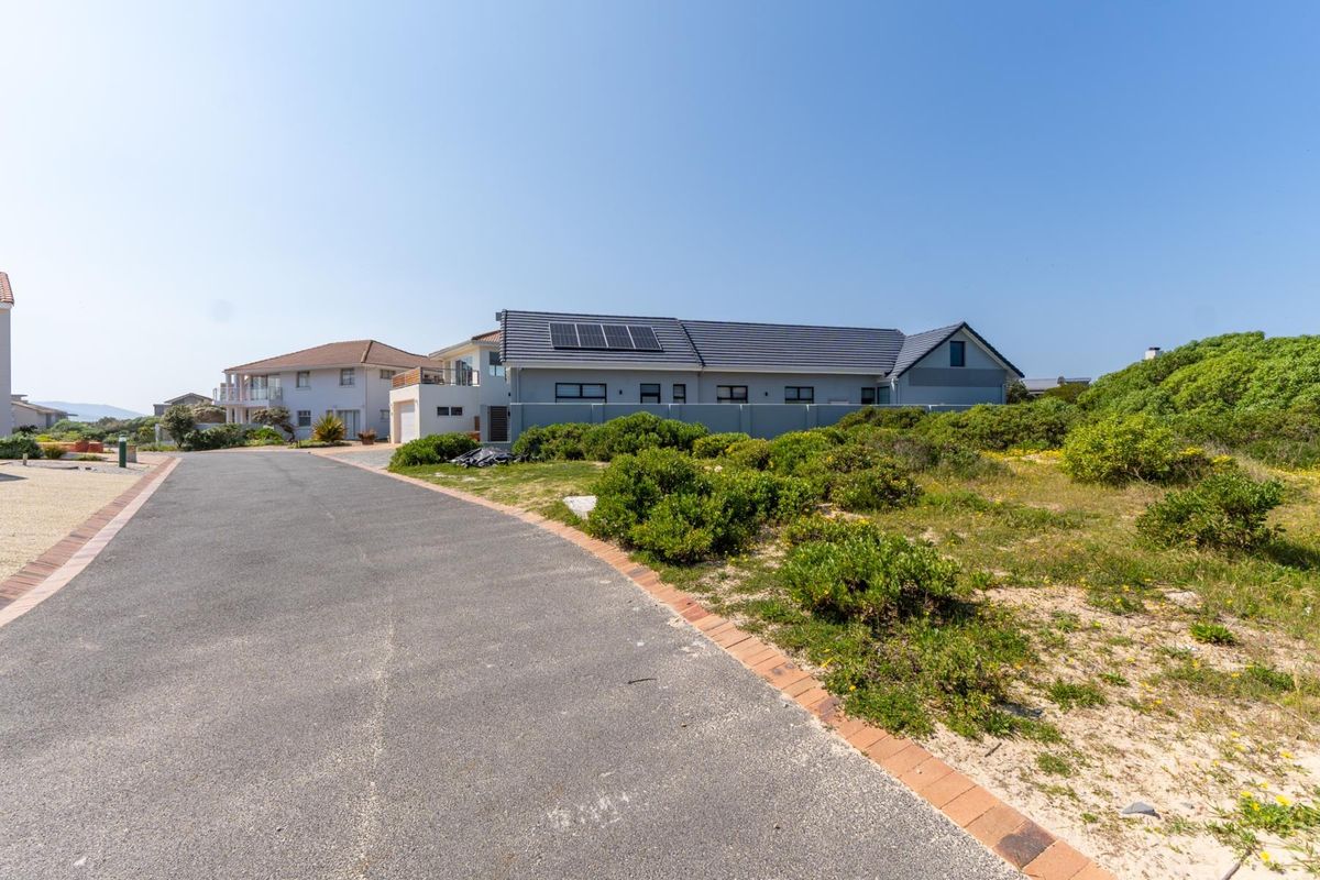 A street view of a residential property featuring a paved road, boundary wall, and a house with solar panels installed on the roof.