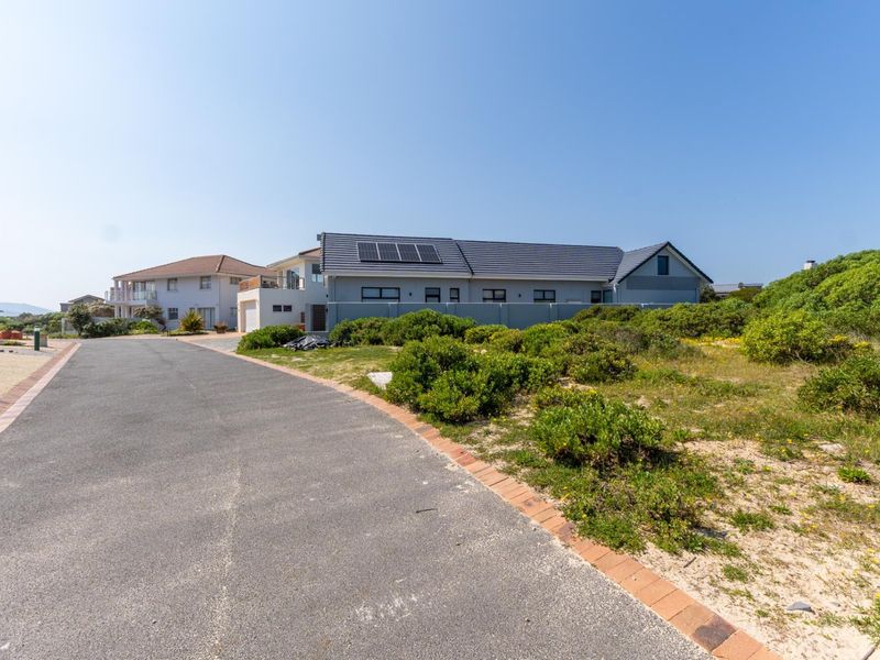 A street view of a residential property featuring a paved road, boundary wall, and a house with solar panels installed on the roof.
