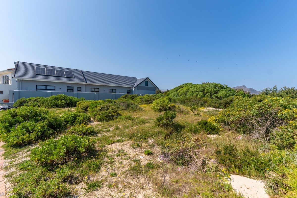 A view of a vacant residential plot featuring natural coastal vegetation, with a neighboring property visible in the background equipped with solar panels.