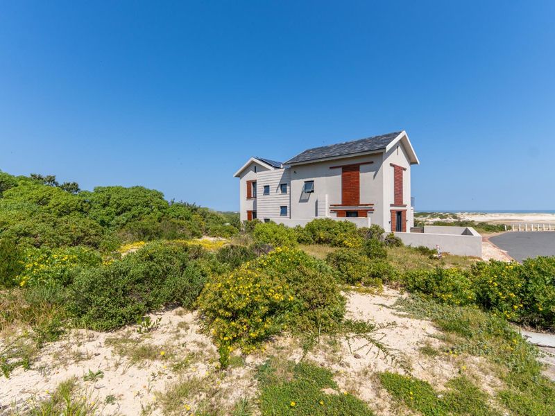 A modern two-story residential property featuring timber shutters and solar panels, situated in a coastal landscape with natural vegetation and a paved driveway.
