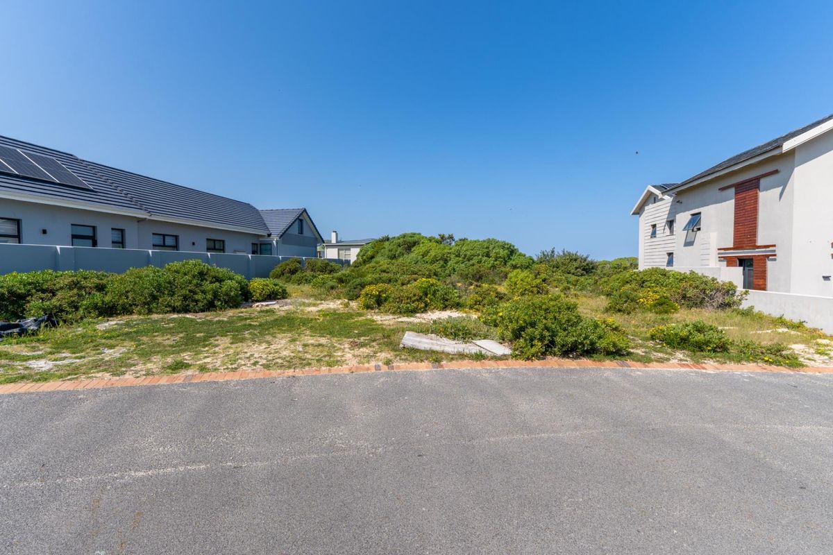 A vacant residential plot situated between two existing modern homes, featuring natural vegetation and direct access from a paved street.