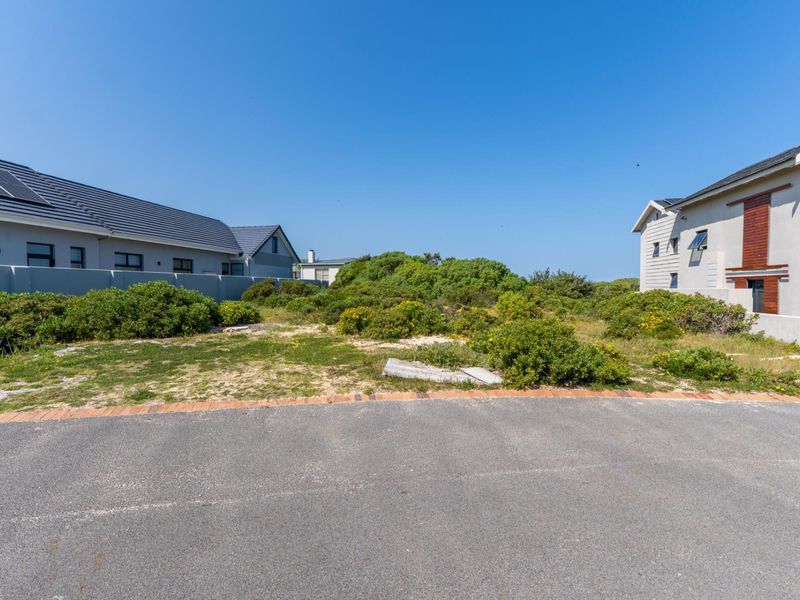 A vacant residential plot situated between two existing modern homes, featuring natural vegetation and direct access from a paved street.