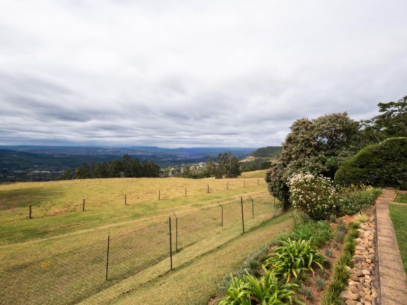Expansive elevated landscape view overlooking rolling hills and valley, featuring a well-maintained garden path and perimeter fencing.