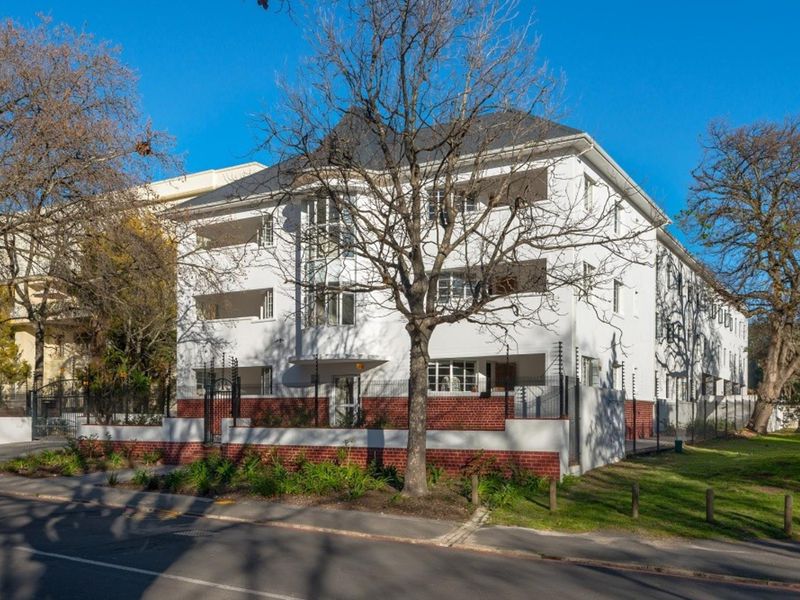 A well-maintained multi-story apartment building featuring a white facade, brick base, and private balconies, secured with perimeter electric fencing.