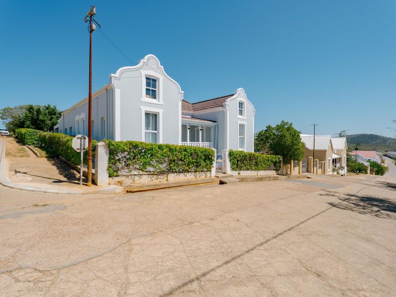 A charming Cape Dutch style house with a gabled facade, sash windows, and a well-maintained hedge boundary, situated on a corner street.