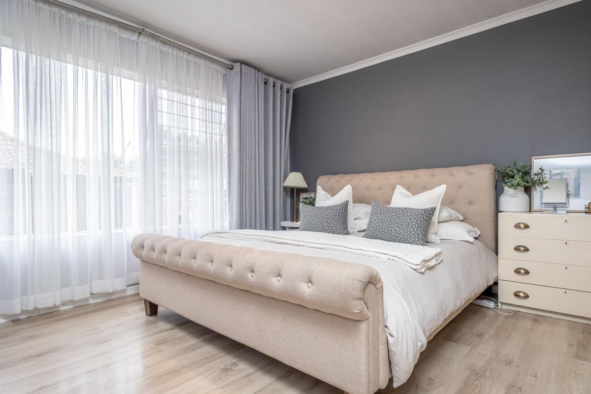 A well-maintained bedroom featuring light-toned laminate flooring, a large window with curtain tracks, and a neutral grey accent wall.