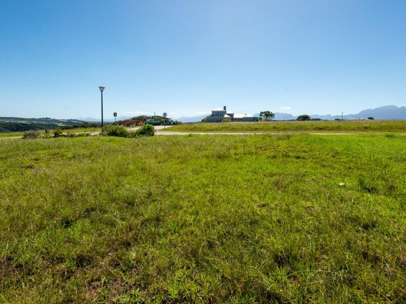 A wide-angle view of a vacant residential plot featuring natural grass coverage, a paved road, and distant mountain views.