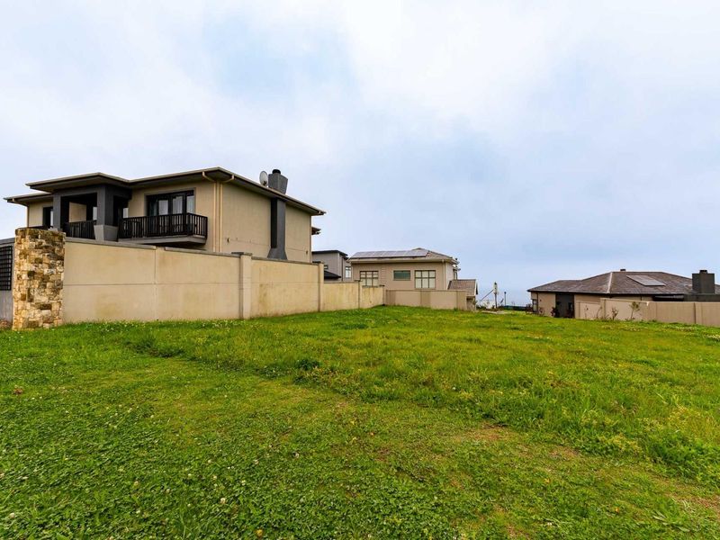 A vacant residential plot featuring a well-maintained grass lawn, enclosed by a boundary wall with stone cladding accents, adjacent to established residential properties.