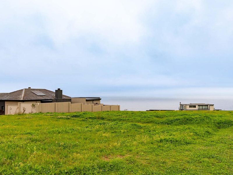 A view of a vacant residential plot featuring grassy terrain with a distant ocean horizon and neighboring properties.