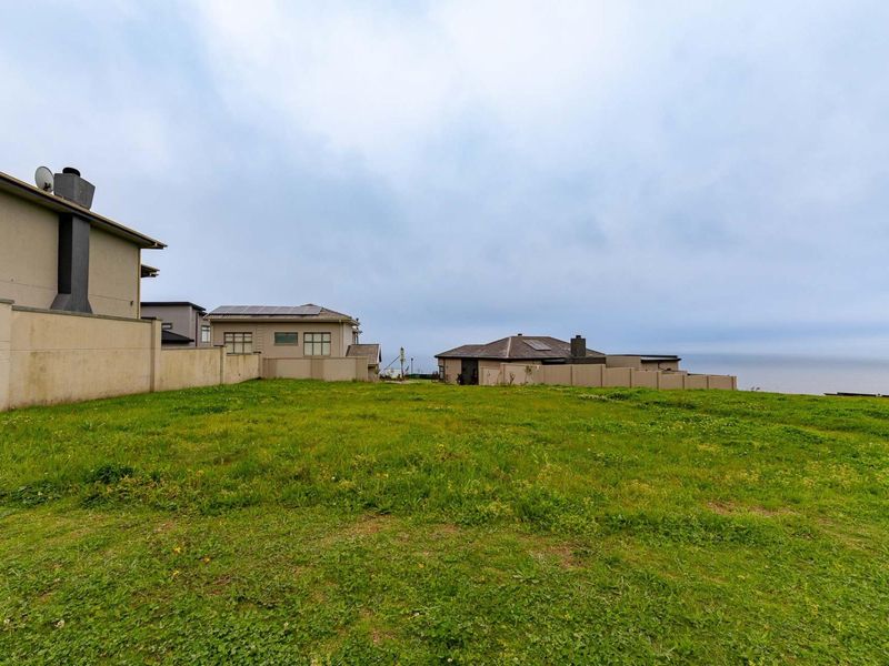 A vacant residential plot featuring a grass lawn and boundary walls, situated in a coastal neighborhood with views of neighboring properties.