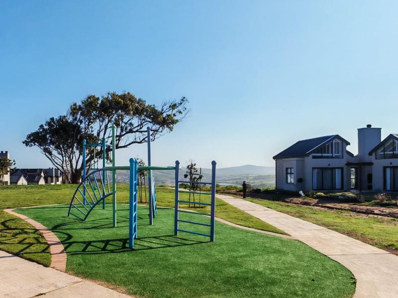 A well-maintained communal outdoor area featuring a playground, paved pathways, and artificial turf, set against a backdrop of residential homes and scenic hills.