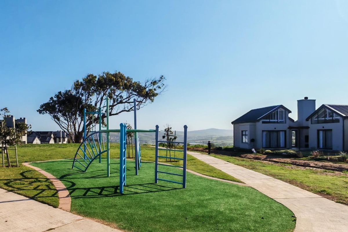 A well-maintained communal outdoor area featuring a playground, paved pathways, and artificial turf, set against a backdrop of residential homes and scenic hills.