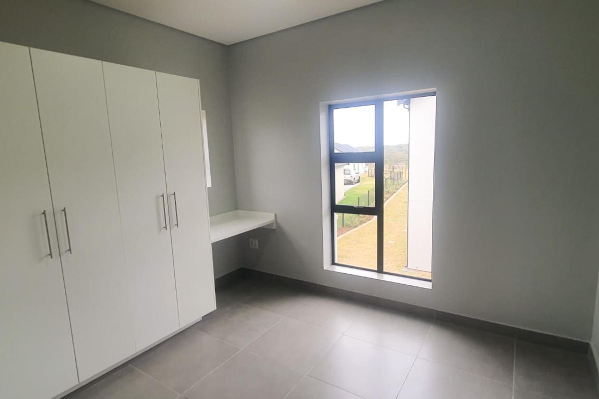 A bright, modern bedroom featuring neutral grey walls, large format floor tiles, built-in cupboards, and a convenient integrated desk space.
