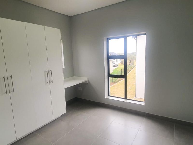 A bright, modern bedroom featuring neutral grey walls, large format floor tiles, built-in cupboards, and a convenient integrated desk space.