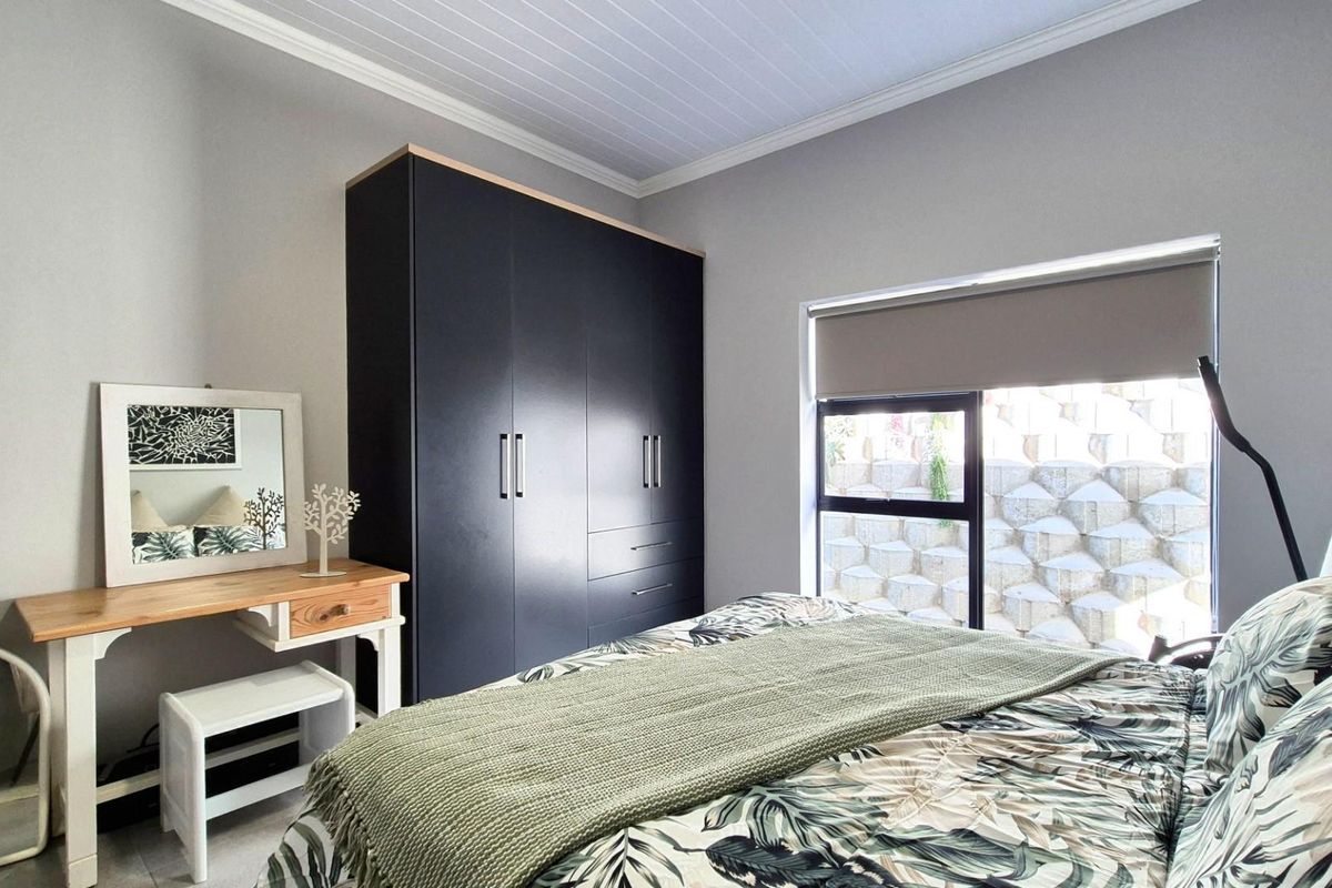 A well-maintained bedroom featuring a modern dark built-in wardrobe, neutral wall finishes, and a window with a roller blind overlooking a retaining wall.