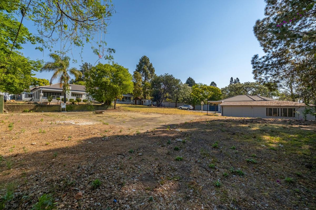 A spacious vacant residential plot featuring mature trees, a paved section, and a retaining wall, offering potential for development or landscaping.