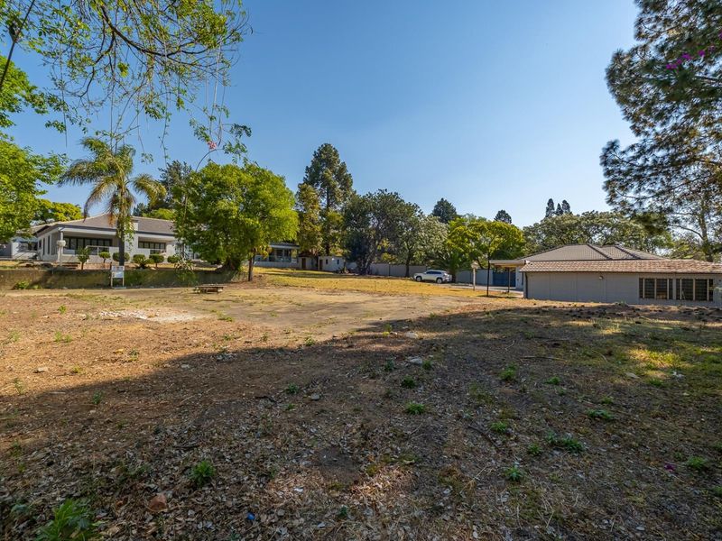 A spacious vacant residential plot featuring mature trees, a paved section, and a retaining wall, offering potential for development or landscaping.