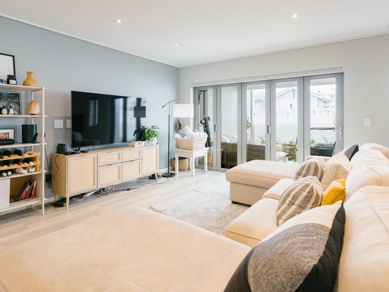 A bright and modern living room featuring light laminate flooring, recessed downlighting, and large aluminium stacker doors providing seamless access to the outdoor patio area.