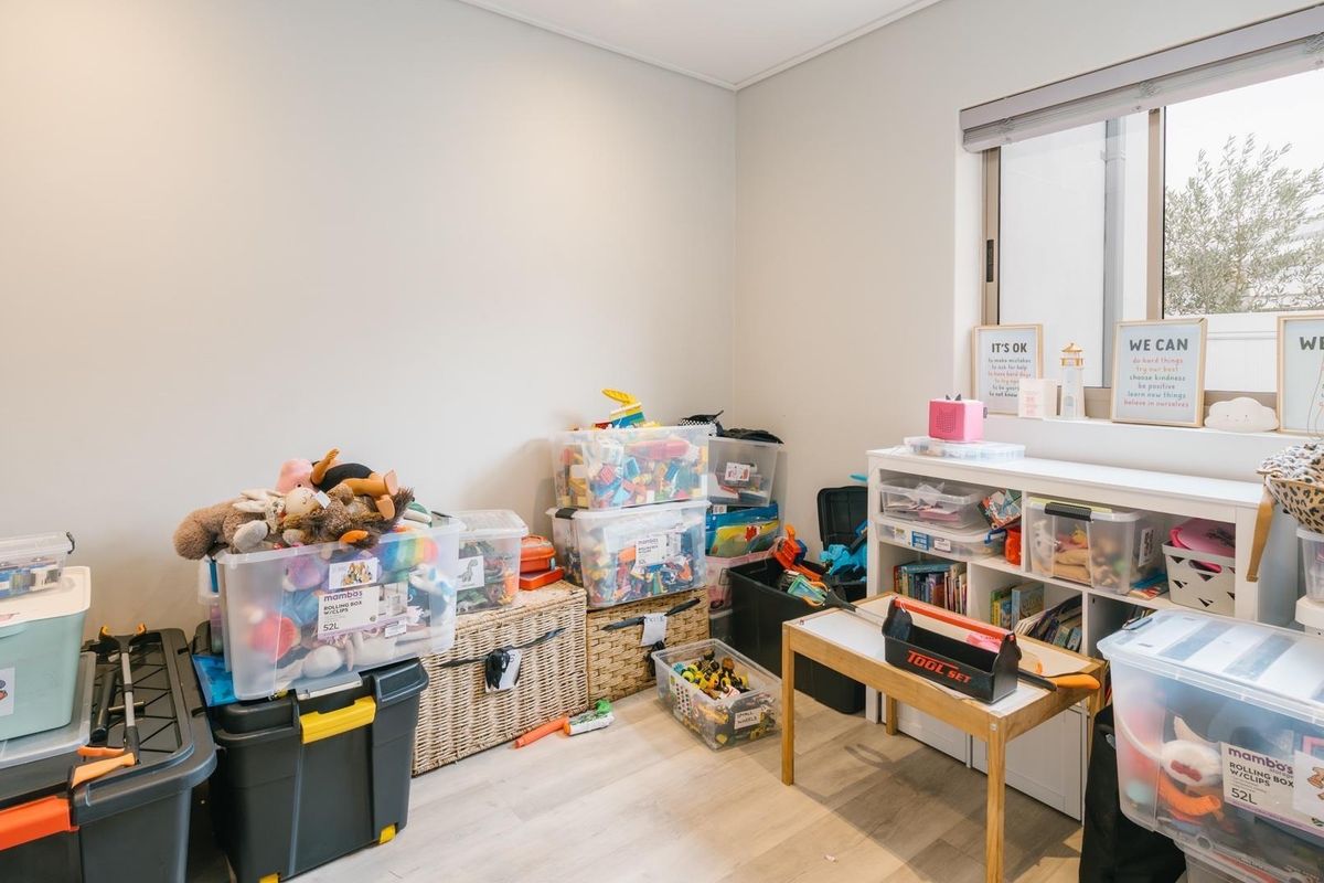 A functional bedroom space featuring light-toned laminate flooring and a window with a fitted roller blind, currently utilized as a play area.