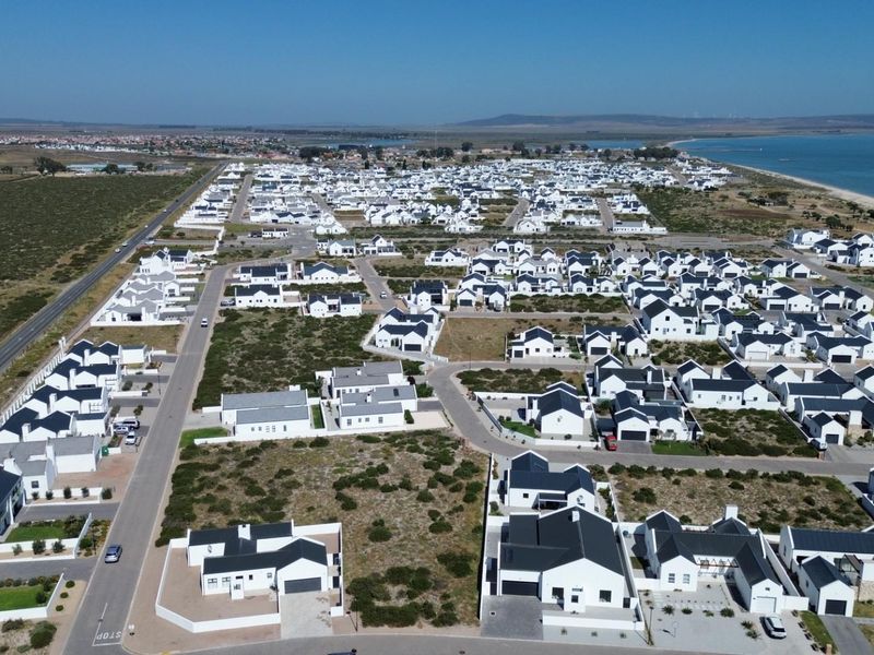 An aerial view of a residential estate featuring uniform white houses with dark roofs, situated near the coastline with established road infrastructure.