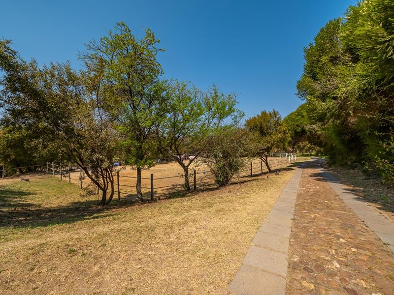 A scenic outdoor area featuring a stone-paved pathway leading through a landscaped garden with mature trees and post and rail fencing.