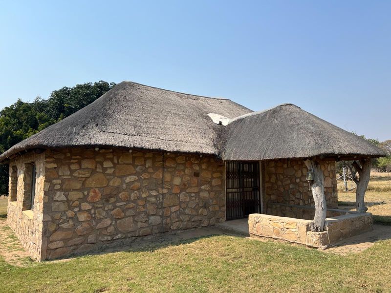 A rustic stone cottage featuring a traditional thatched roof and a covered front porch area, set within a rural farm landscape.