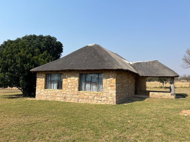 A charming stone-clad farmhouse featuring a traditional thatched roof and a covered patio area, set within a spacious rural landscape.