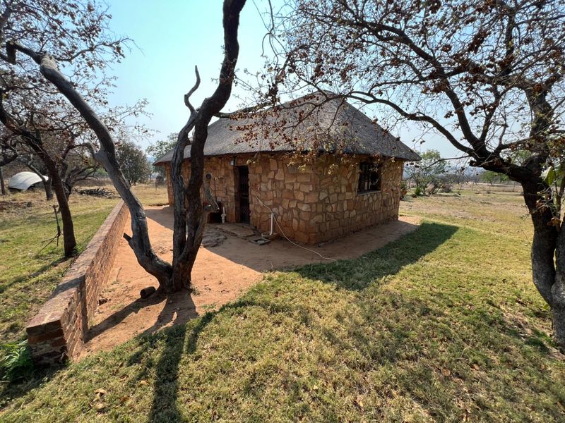 A rustic stone-clad cottage with a traditional thatch roof, situated in a natural farm landscape with a low brick retaining wall.