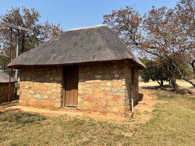 A rustic stone-built cottage with a traditional thatched roof, featuring an integrated solar panel setup in a rural farm setting.