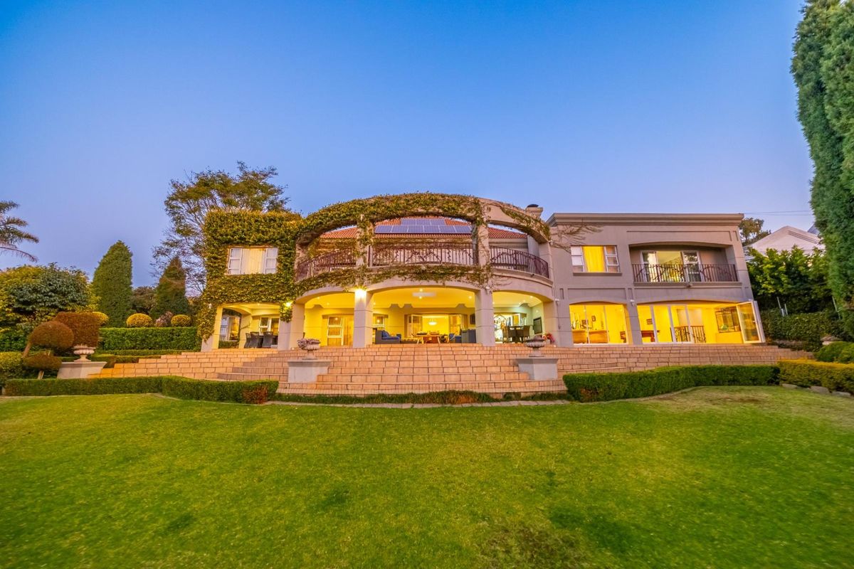 Spacious rear elevation featuring a large covered patio, upper balcony with wrought iron railings, stone staircase, and expansive landscaped garden.