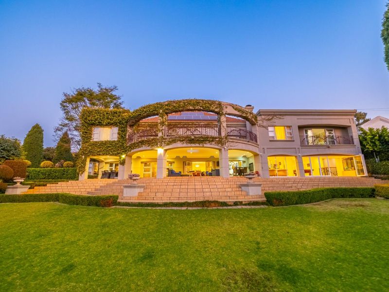 Spacious rear elevation featuring a large covered patio, upper balcony with wrought iron railings, stone staircase, and expansive landscaped garden.