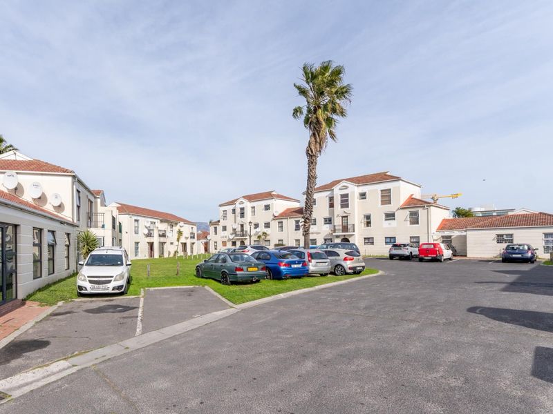 Exterior view of an apartment complex featuring a paved parking area, landscaped lawn, and multi-story residential buildings with tiled roofs.