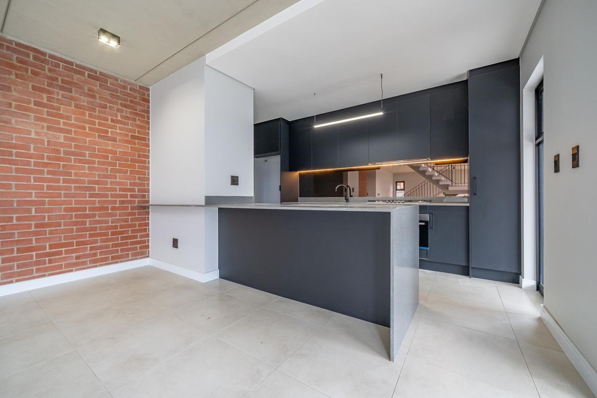 Modern kitchen featuring dark cabinetry, stone countertops, a central island, and an exposed brick feature wall, finished with sleek tiled flooring and recessed lighting.