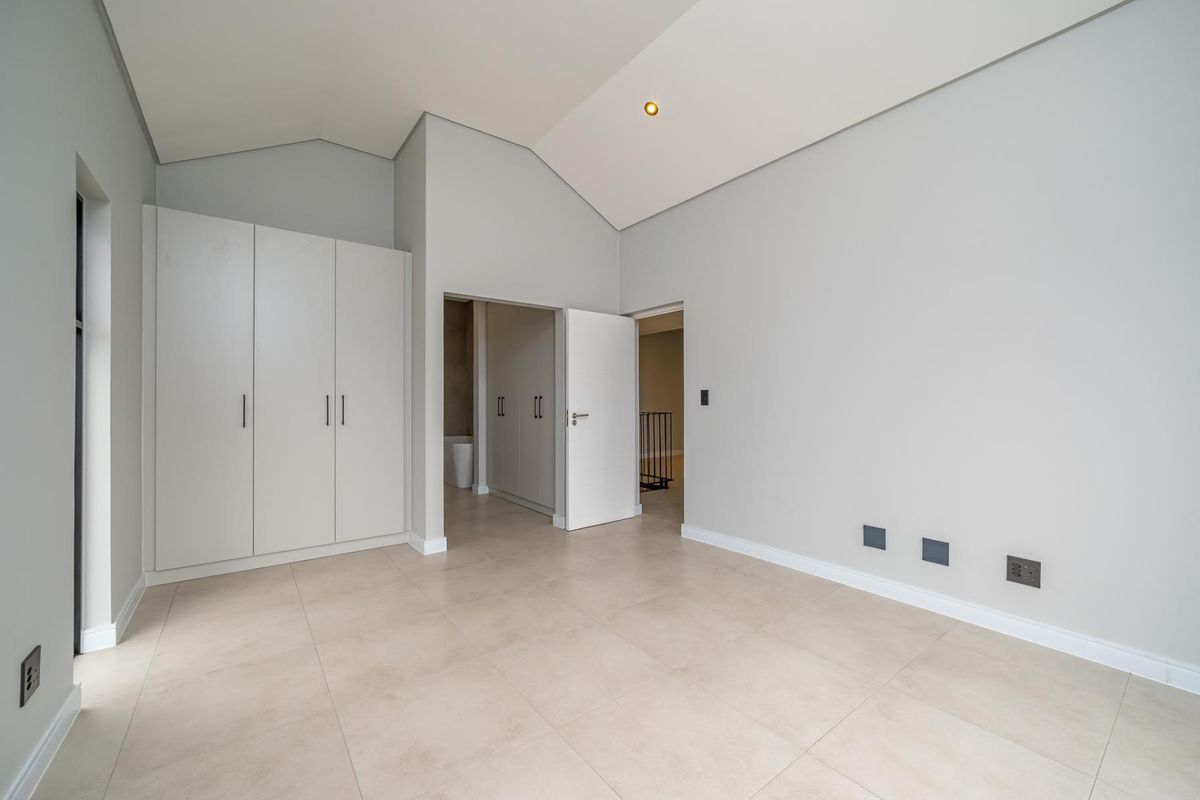 A bright, modern bedroom featuring vaulted ceilings, neutral tiled flooring, and ample built-in cupboard space, leading into an adjoining en-suite area.